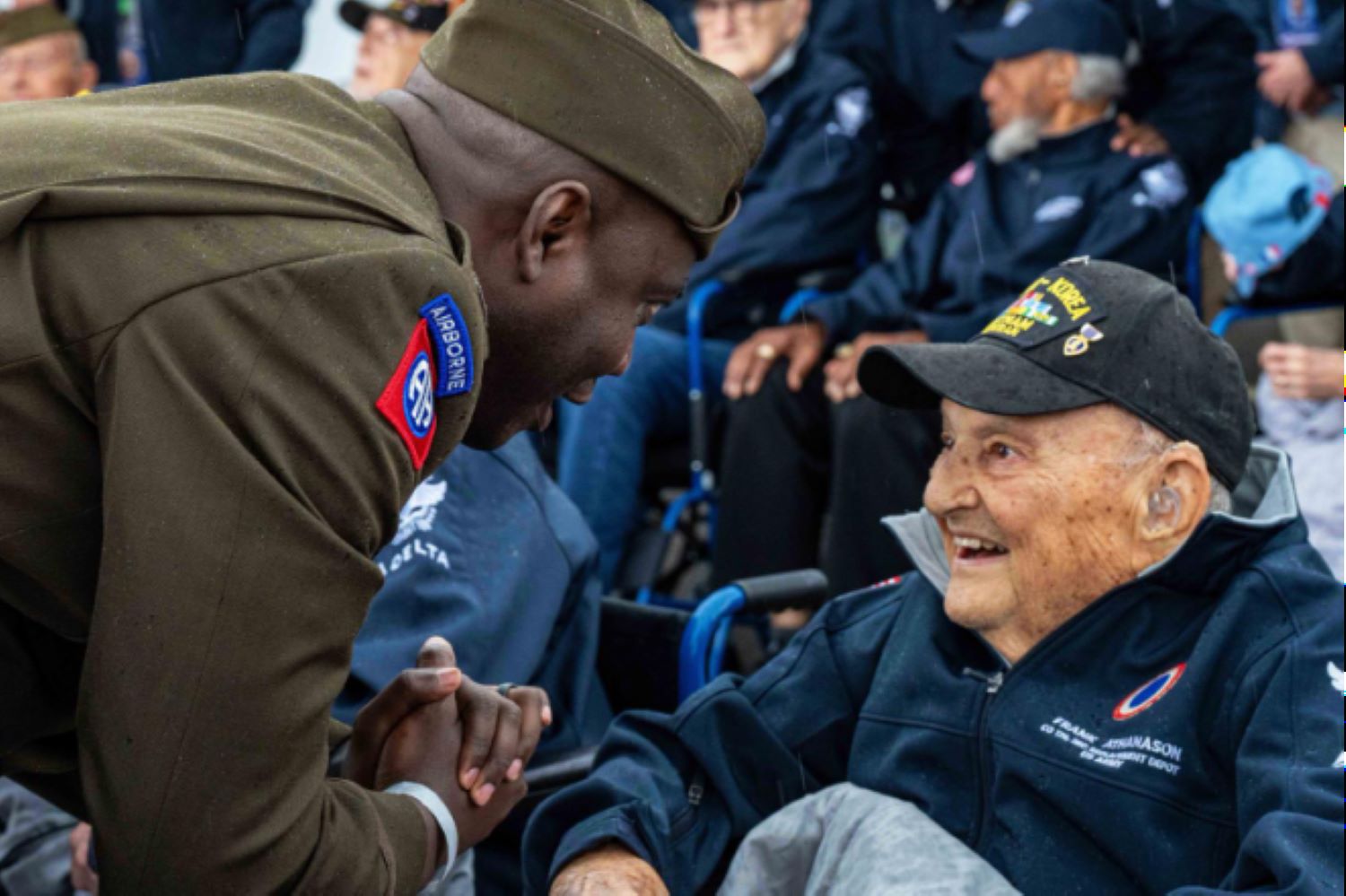 A U.S. Army soldier from the 82nd Airborne Division leans forward with a warm smile, clasping hands with a World War II veteran seated in a wheelchair.
